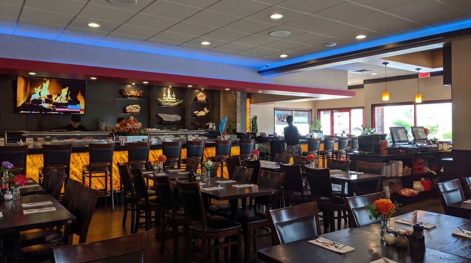 Empty restaurant dining room with dark tables and chairs, warm lighting, and a bar area in the background