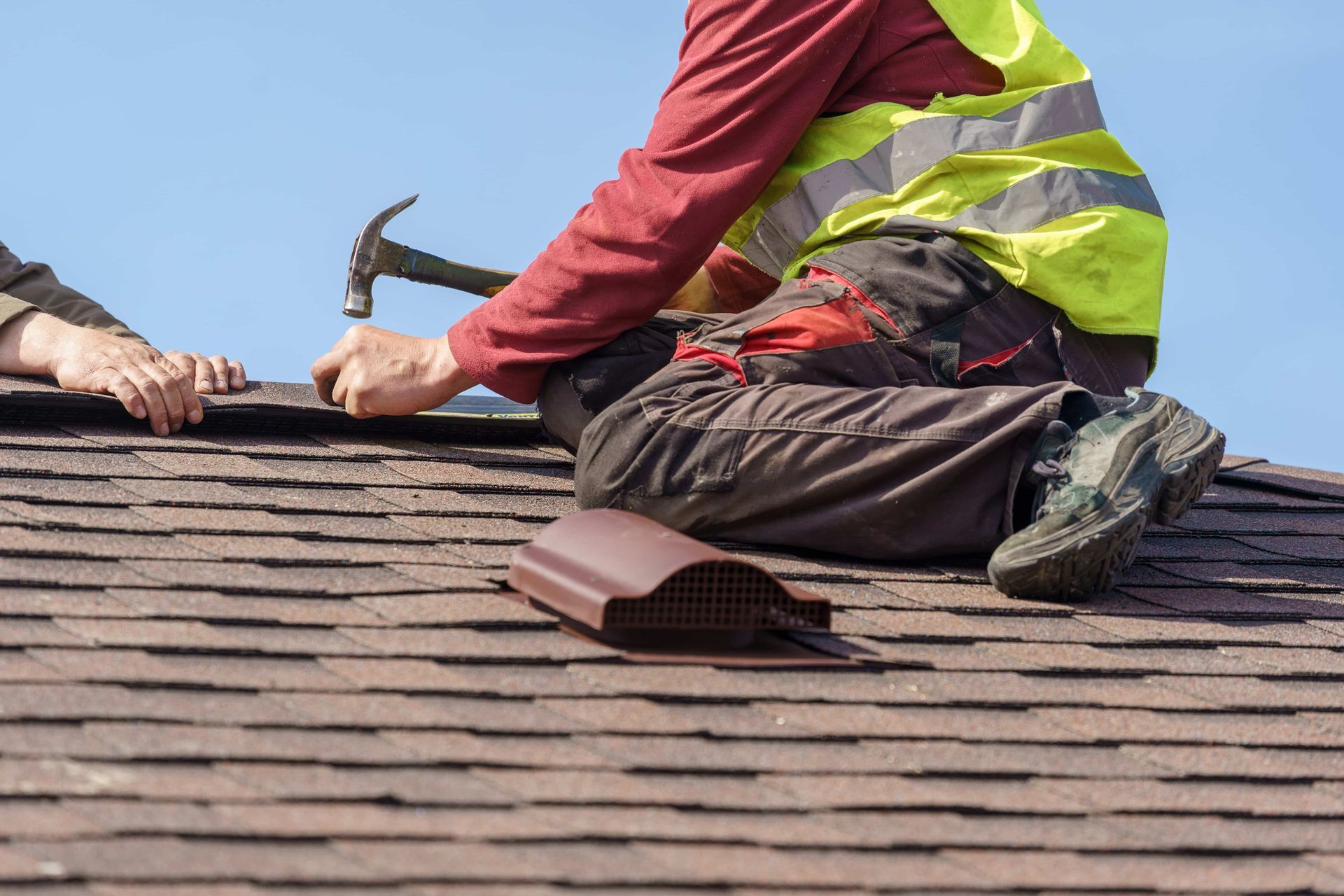 Roofing worker kneeling on shingles, hammering near a skylight under a blue sky