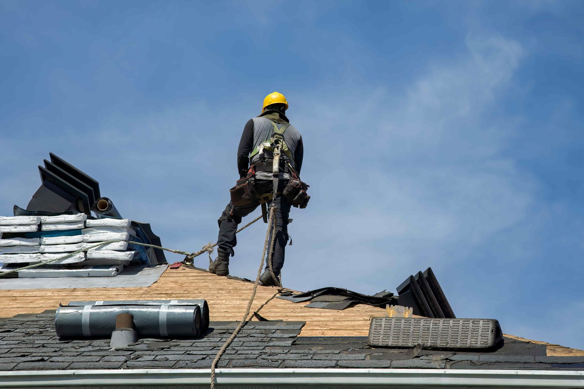 Worker in yellow hard hat standing on a roof, repairing shingles under a blue sky.