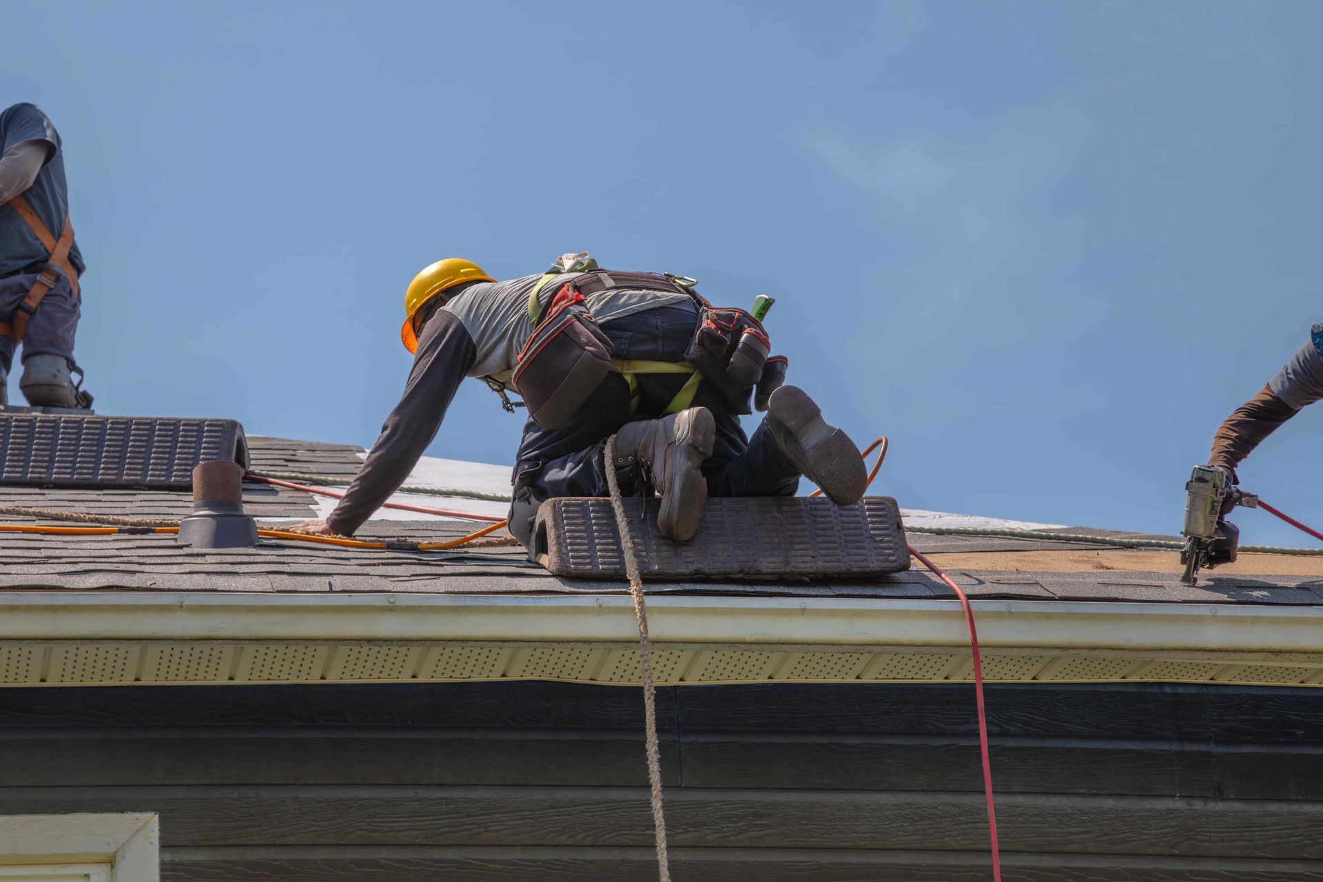 Roofers working on a rooftop, one kneeling near a harness rope under a clear sky