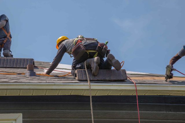 Roof workers crouched on a shingled roof, securing equipment under a clear blue sky