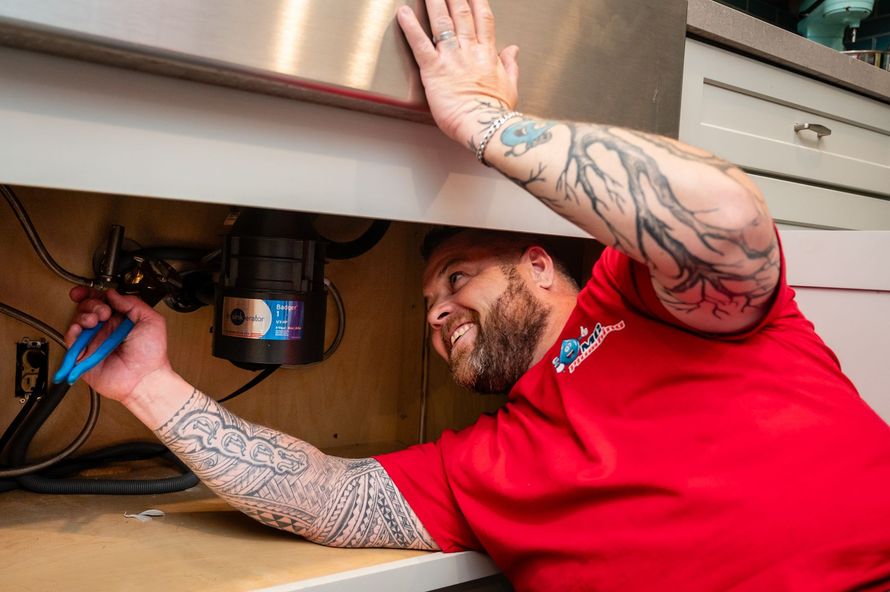 Person with tattoos repairing plumbing under a sink with a blue tool, wearing a red shirt and smiling