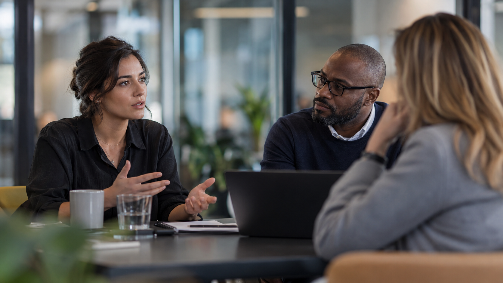 Three coworkers in discussion at a conference table with a laptop and mugs in a modern office
