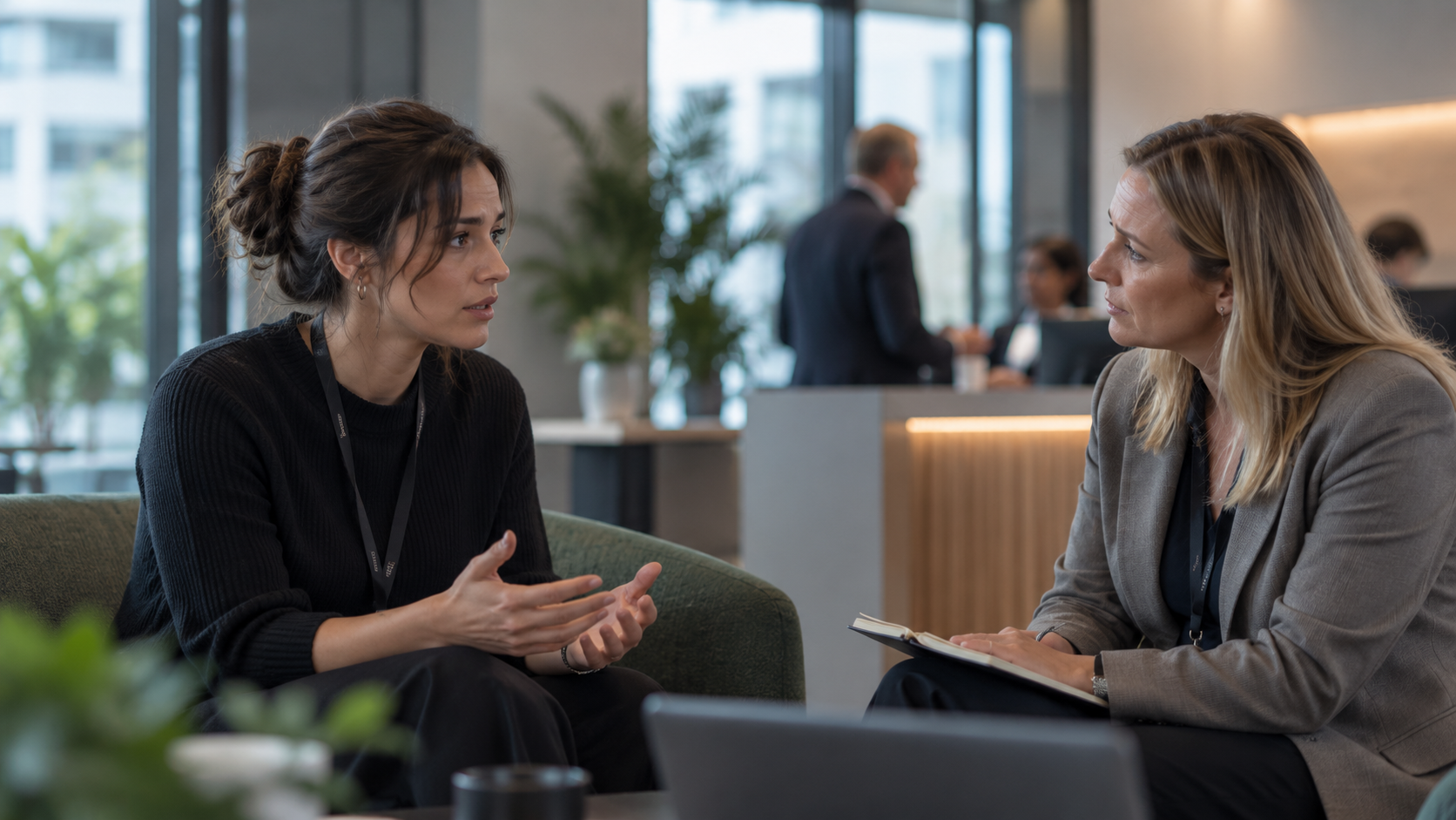 Two people talking in a modern office lounge, seated across from each other and holding papers.