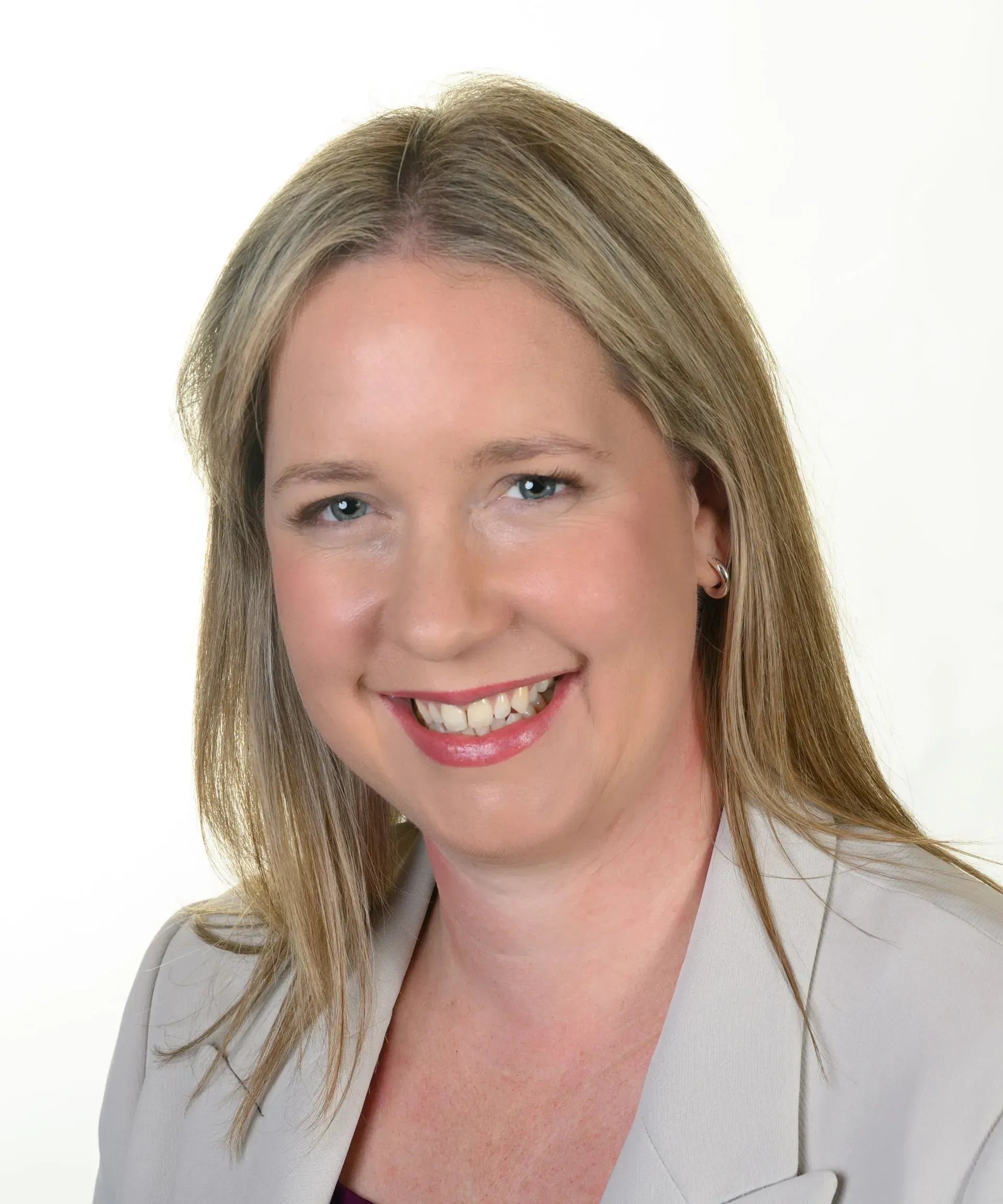 A professional headshot of a smiling person with shoulder-length blonde hair, wearing a light grey blazer.