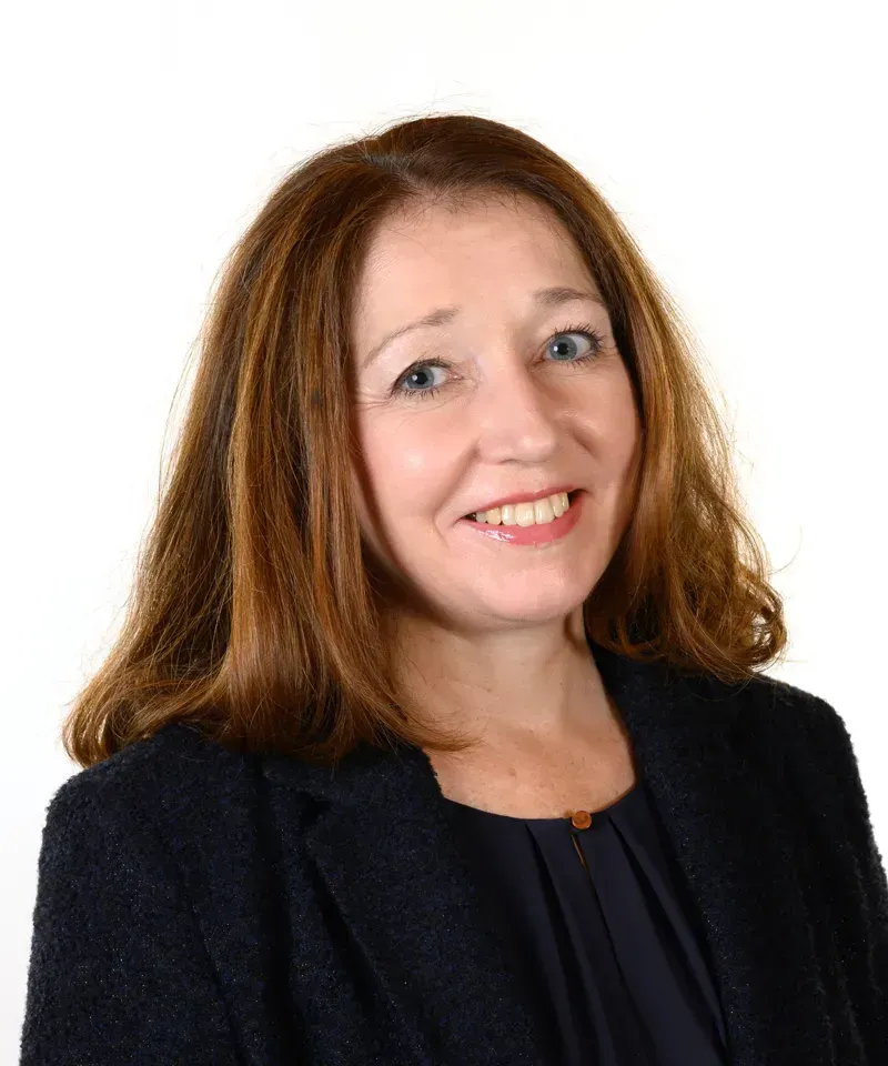 A professional headshot of a smiling person with shoulder-length brown hair, wearing a dark blue blazer over a dark top.