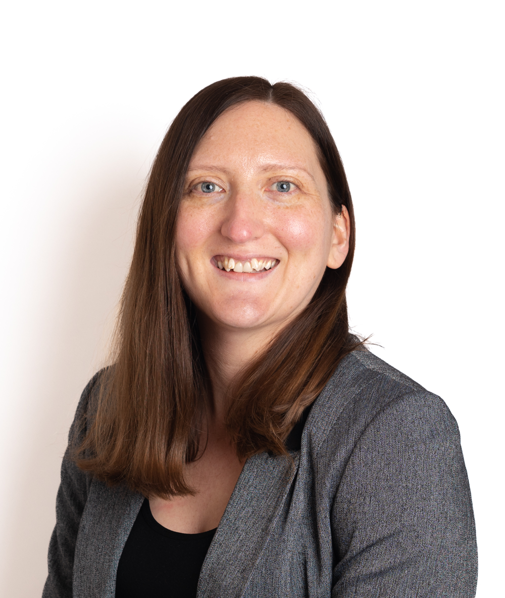 A person with long brown hair smiling against a plain white background, wearing a gray blazer over a black top.