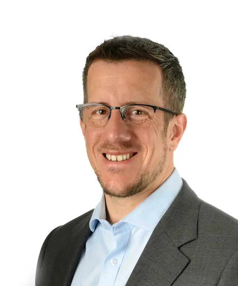 A professional headshot of a person with short brown hair, wearing glasses, a light blue collared shirt, and a gray blazer.