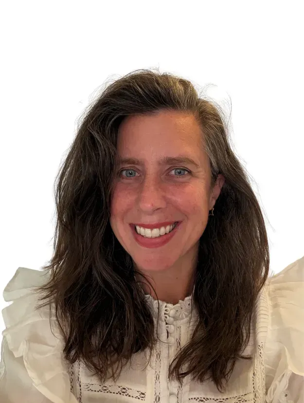 A person with shoulder-length wavy brown hair smiling at the camera, wearing a white blouse with ruffle details.