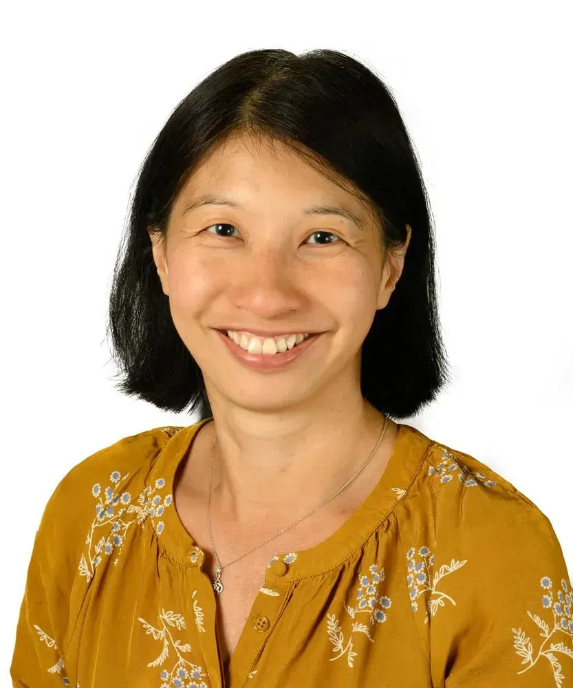 A person with dark hair smiling while wearing a yellow floral blouse against a white background.