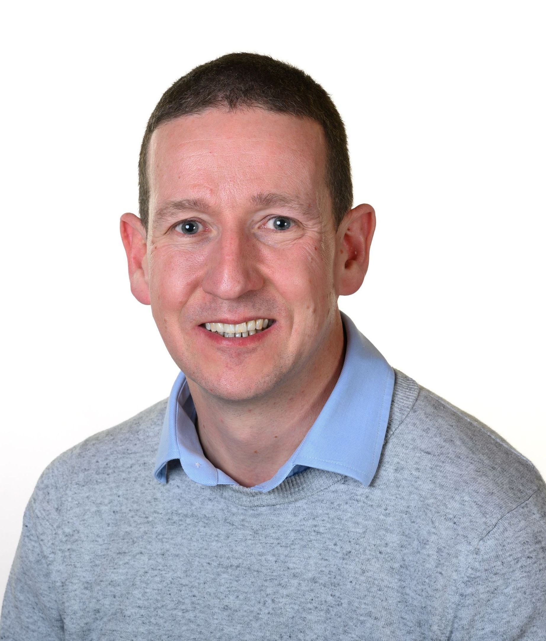 A smiling person wearing a light blue collared shirt and a gray textured sweater against a plain white background.