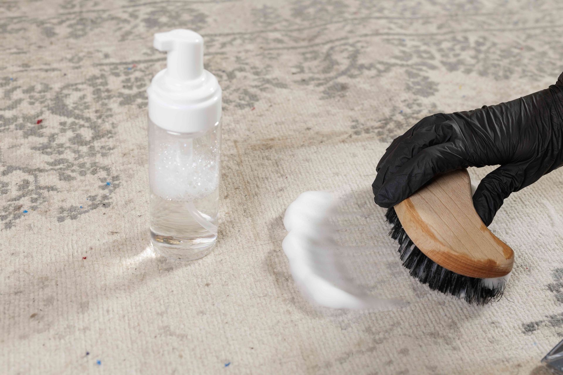 Gloved hand scrubbing a carpet stain with a brush beside a spray cleaner bottle