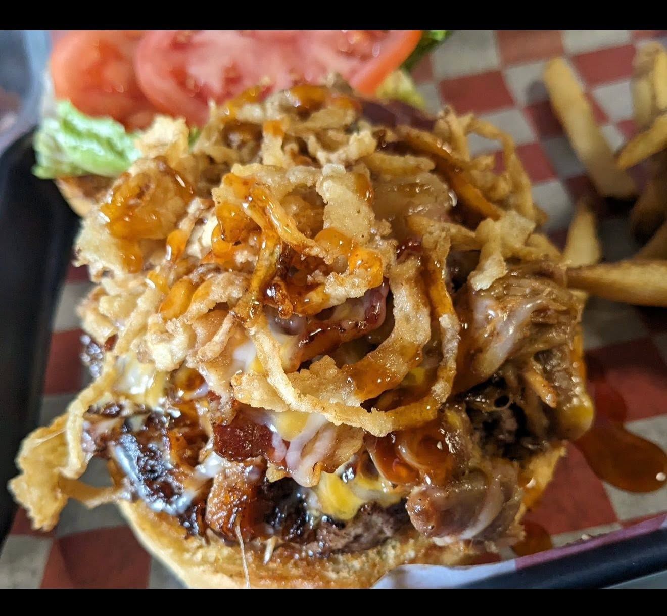 Loaded cheeseburger topped with onion straws and barbecue sauce, with fries and tomato slices on a tray.