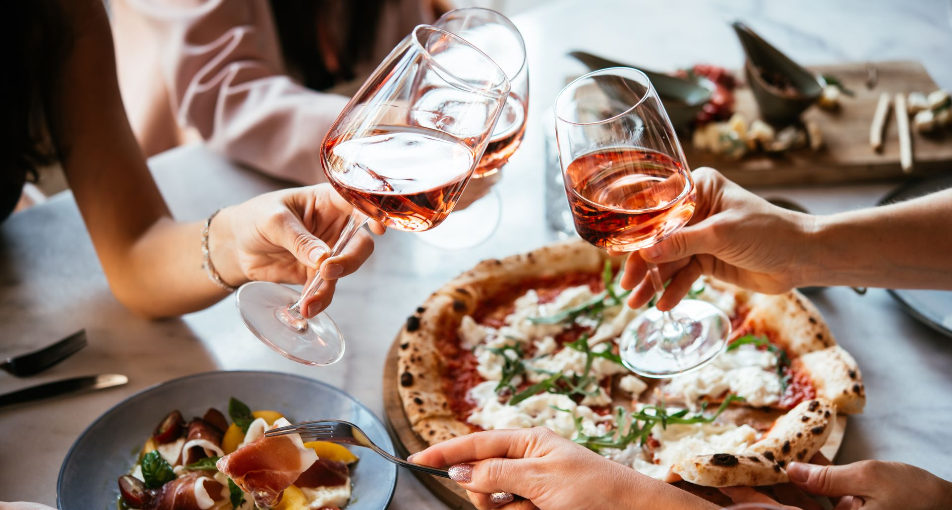 Friends clink wine glasses over pizza and salad at a casual dinner table