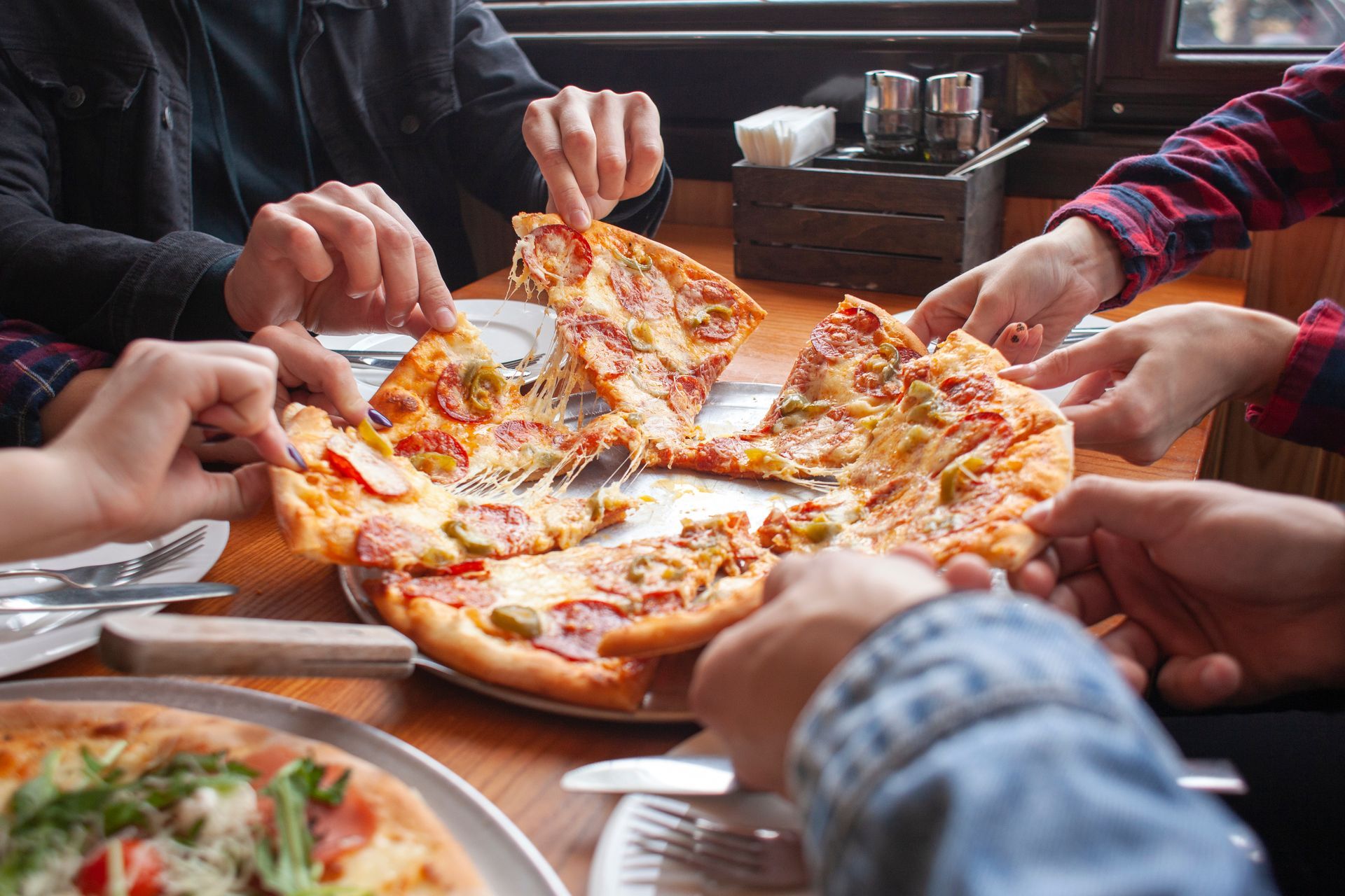 People reaching for slices of pizza on a wooden table in a restaurant.