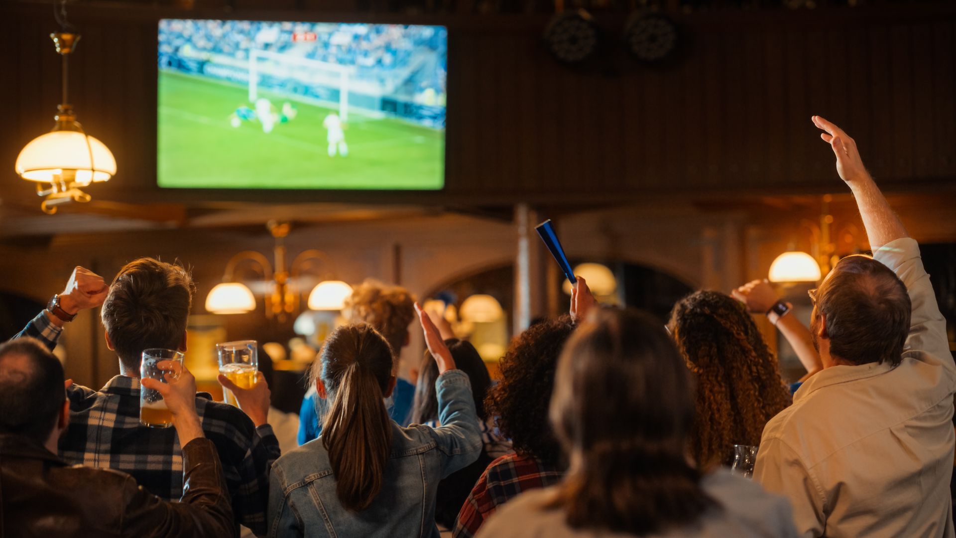 Crowd cheering in a dim bar while watching a soccer match on a large TV screen