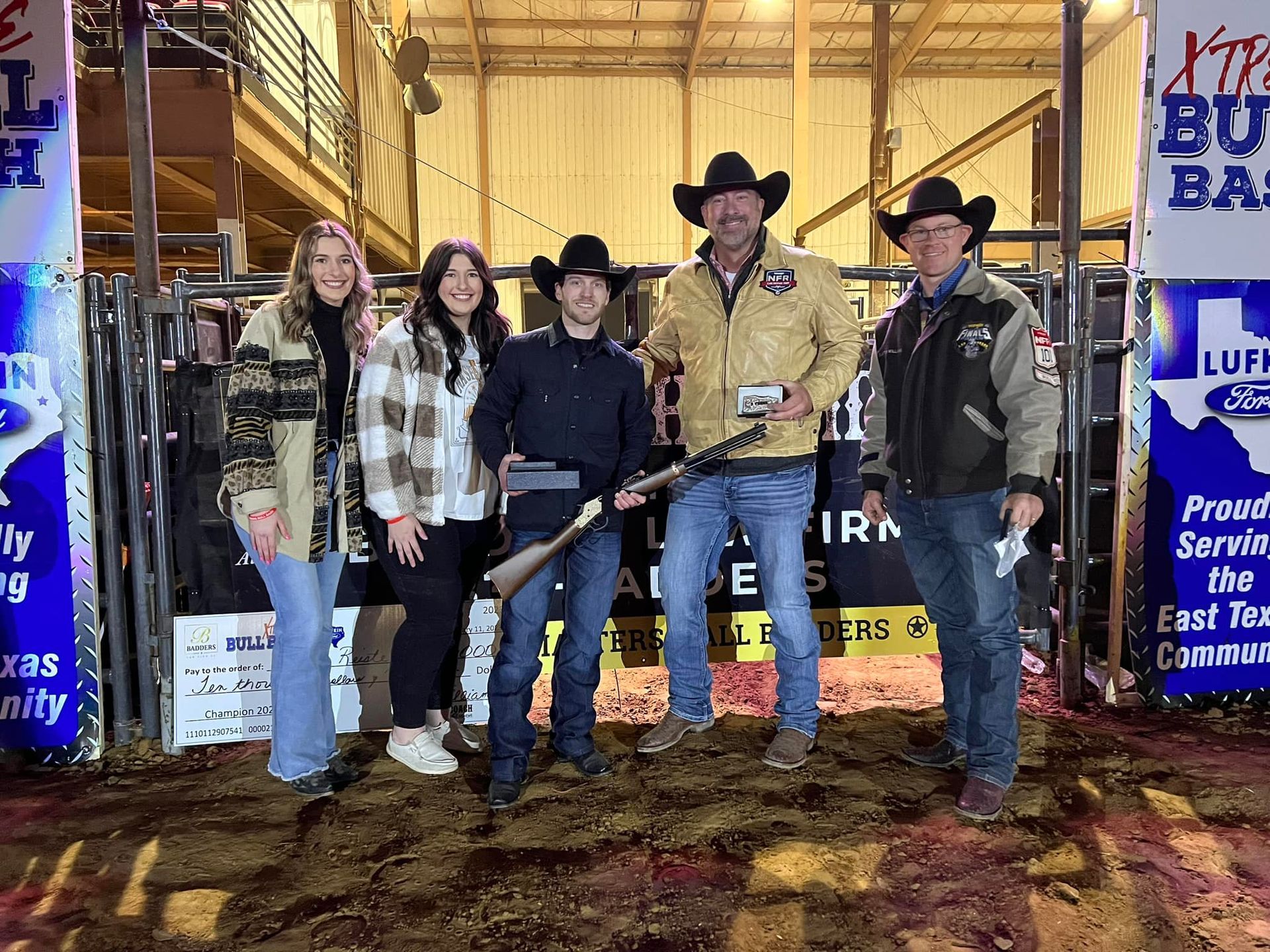 Group of people posing with a rifle at a rodeo event. They're in front of a bull riding arena.