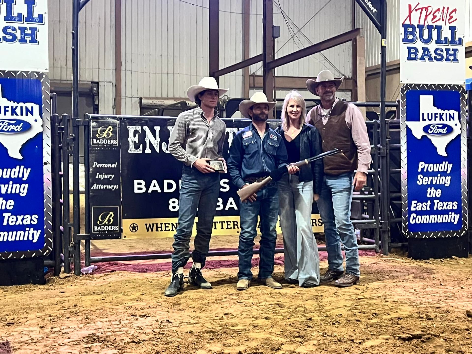 Four people posing at a bull riding event: two men wearing cowboy hats, and two women. Event banner in the background.