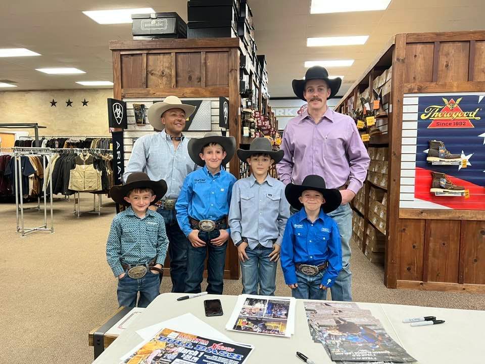 Group of cowboys posing in a Western store, some with belt buckles, smiling. Behind a table with autographs.