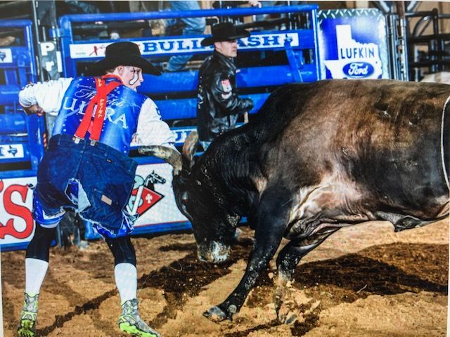 Bull rider in vest, jeans, and boots prepares for a bull in an arena with sponsor signage.