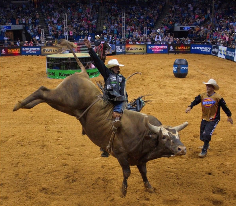 Bull rider in arena, riding a bucking bull, arms raised. Another man in the background.