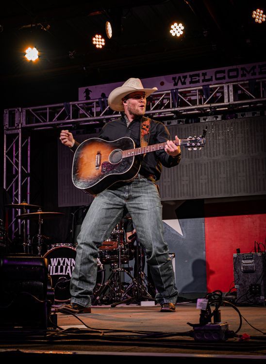 Man in cowboy hat plays acoustic guitar on stage, singing.