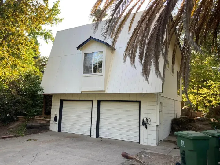 White two-story building with garage doors, an upstairs window, and surrounding trees. Green trash bins are nearby.