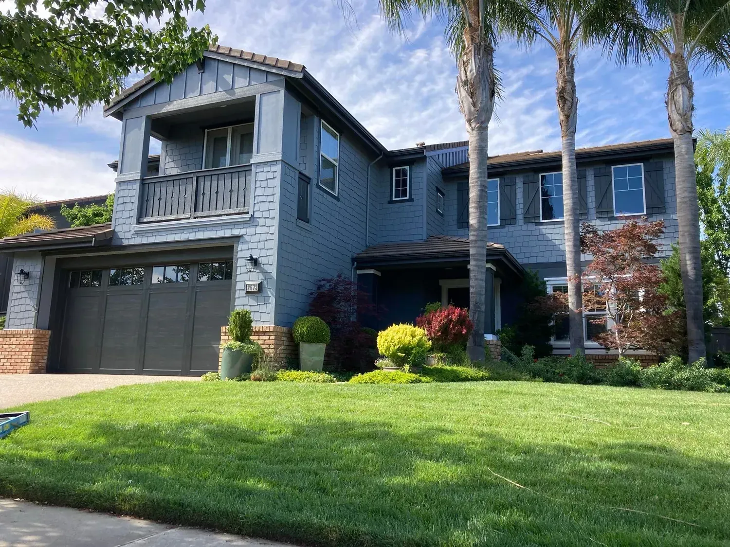 Two-story blue house with a green lawn, palm trees, and shrubs.