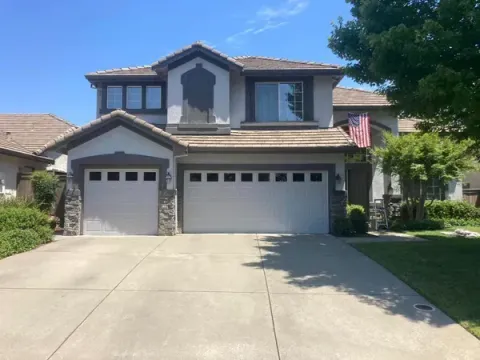 Two-story house with a concrete driveway. Garage doors, stone accents, and an American flag.