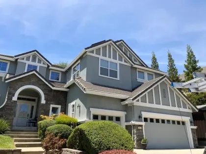 Two-story house with blue-gray stucco, stone accents, white trim, and a dark roof. Green bushes and a blue sky.