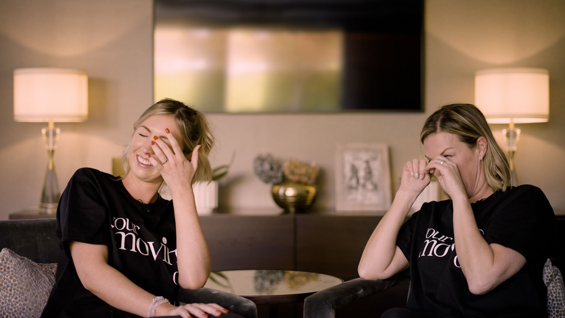Two women laughing, seated in a living room, both wiping their eyes.