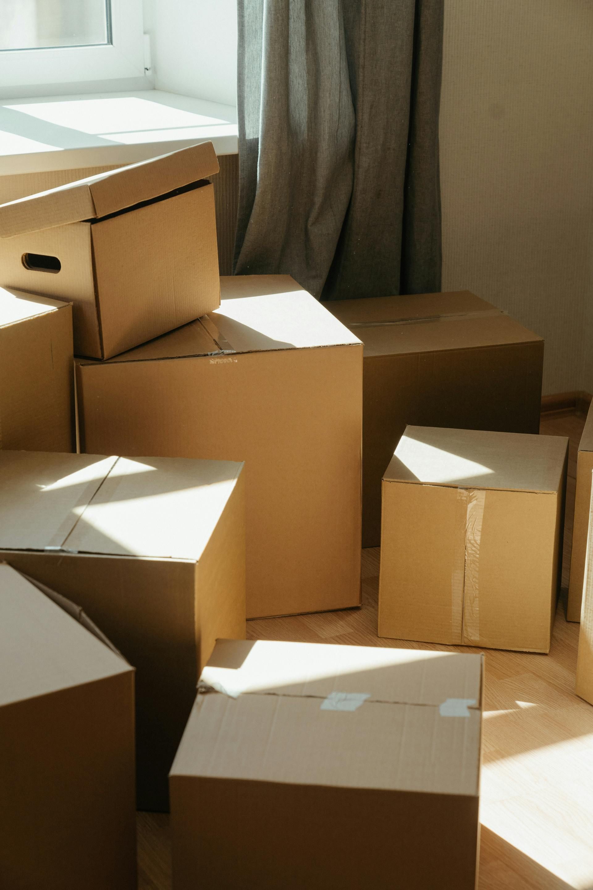 Cardboard moving boxes stacked in a room, sunlight streaming in from a window with a curtain.