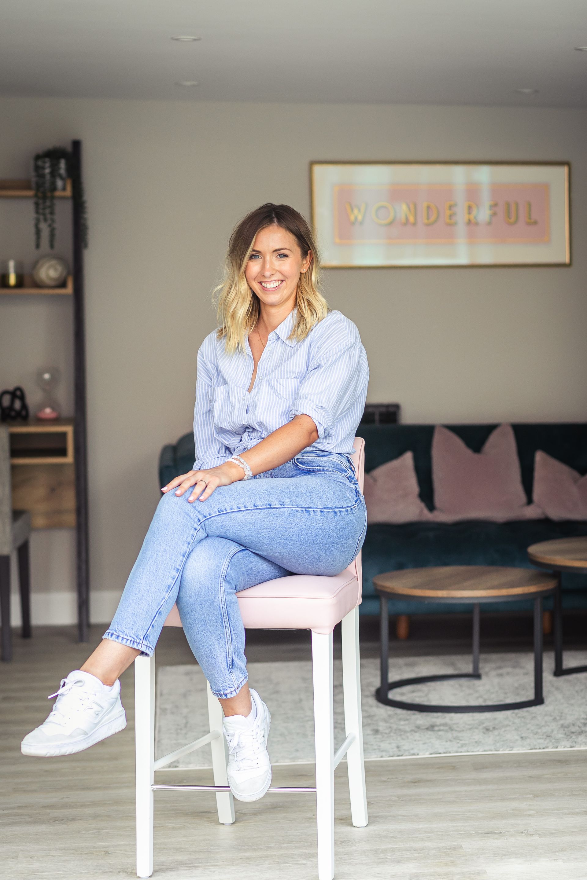 Woman sitting on a pink stool, smiling. Wearing a blue shirt and jeans, white sneakers. Interior setting.