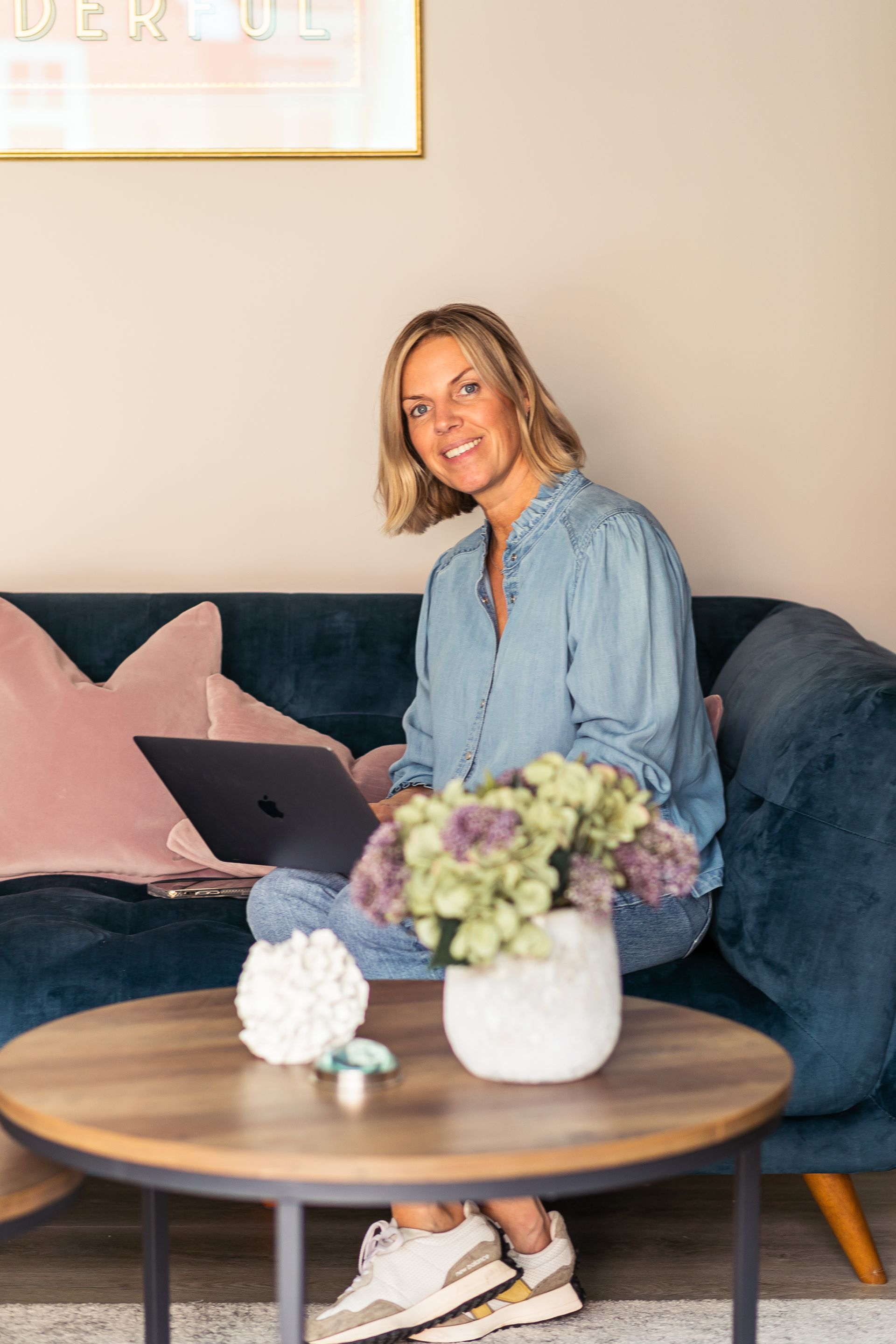 Woman using laptop on a teal couch, smiling, in a home setting. A coffee table with flowers is in front.