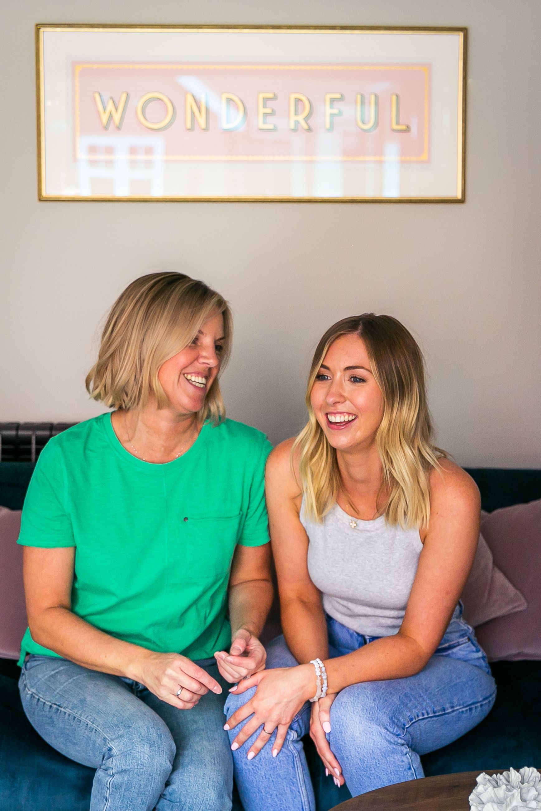 Two women laugh on a teal couch, a