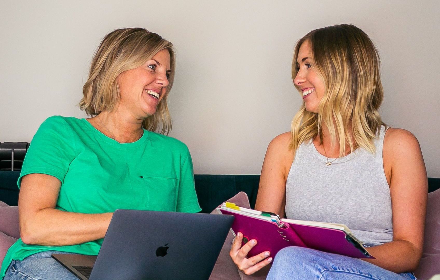 Two women smiling, working together on a laptop and notebook. One wears green, the other a grey tank.