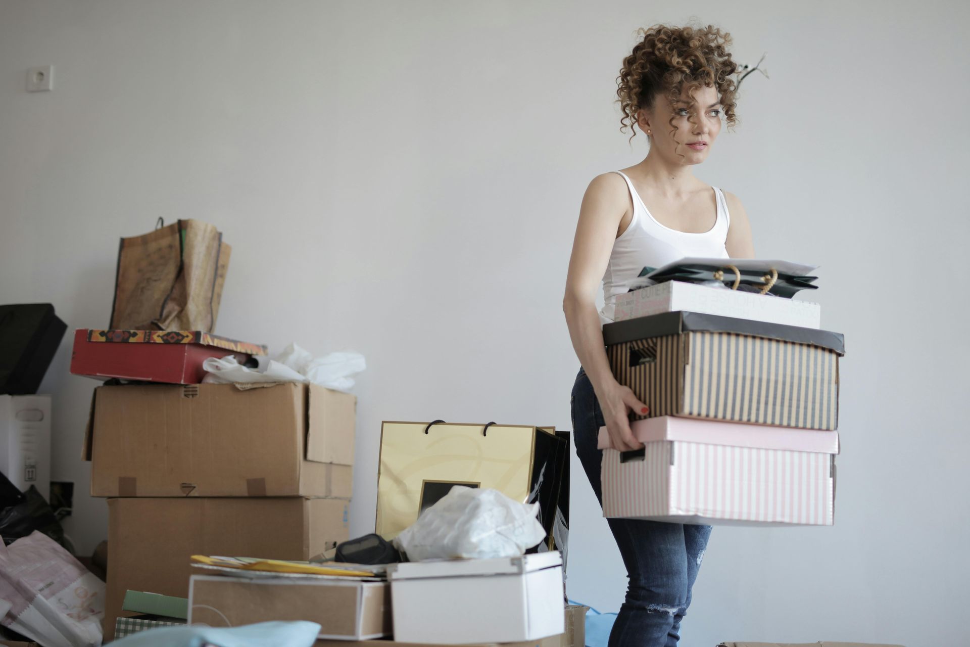 Woman holding stack of boxes indoors, looking stressed; other boxes on the floor.