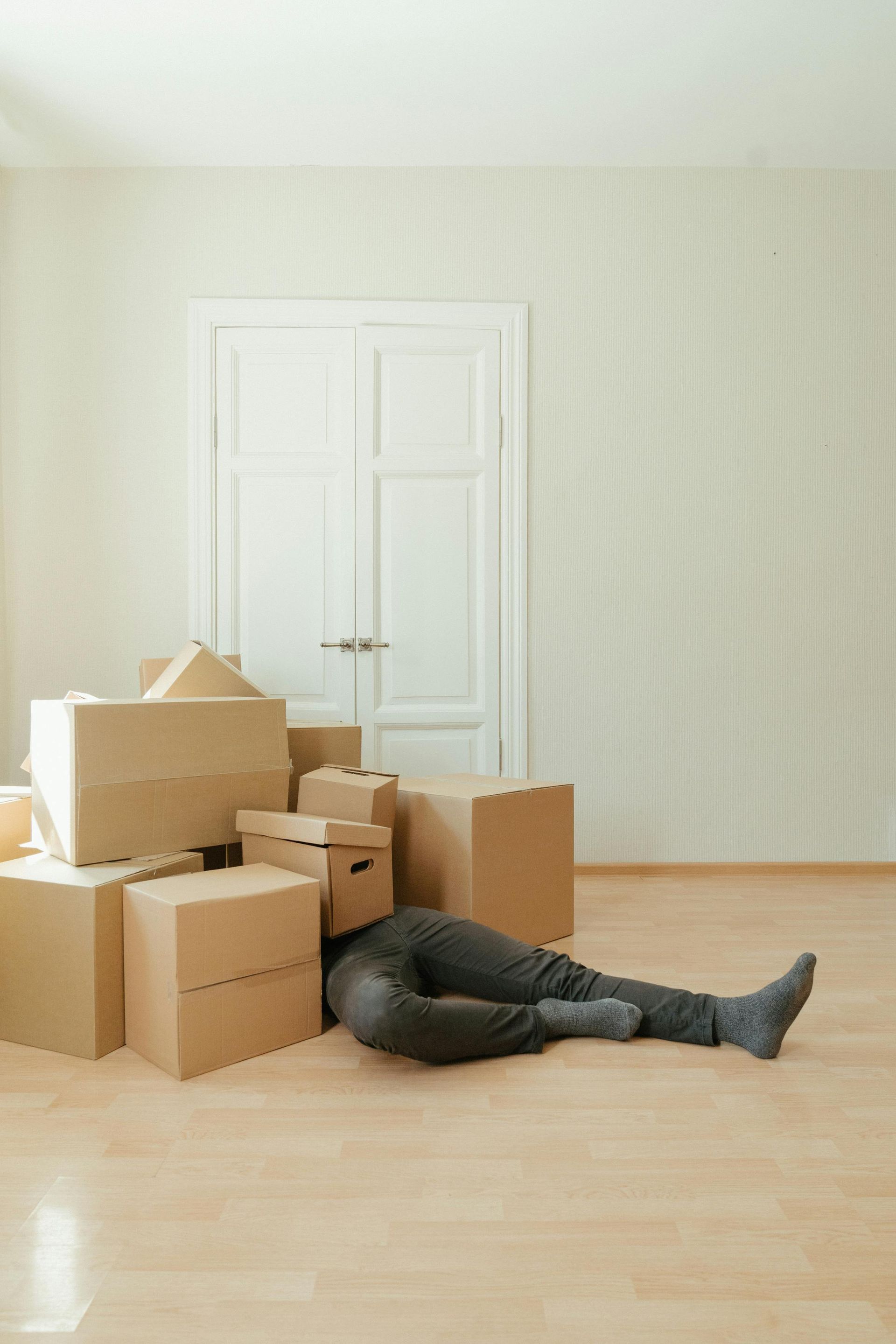Person lying on floor with head in a box, surrounded by moving boxes in a room.