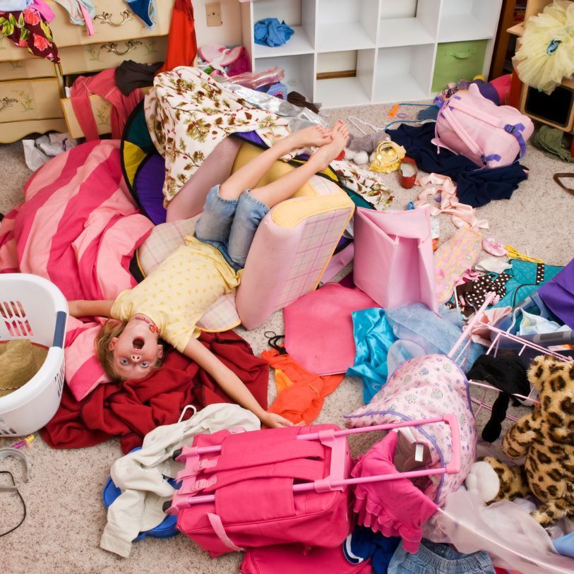 Girl lying in messy room, surrounded by clothes, toys, and a laundry basket.