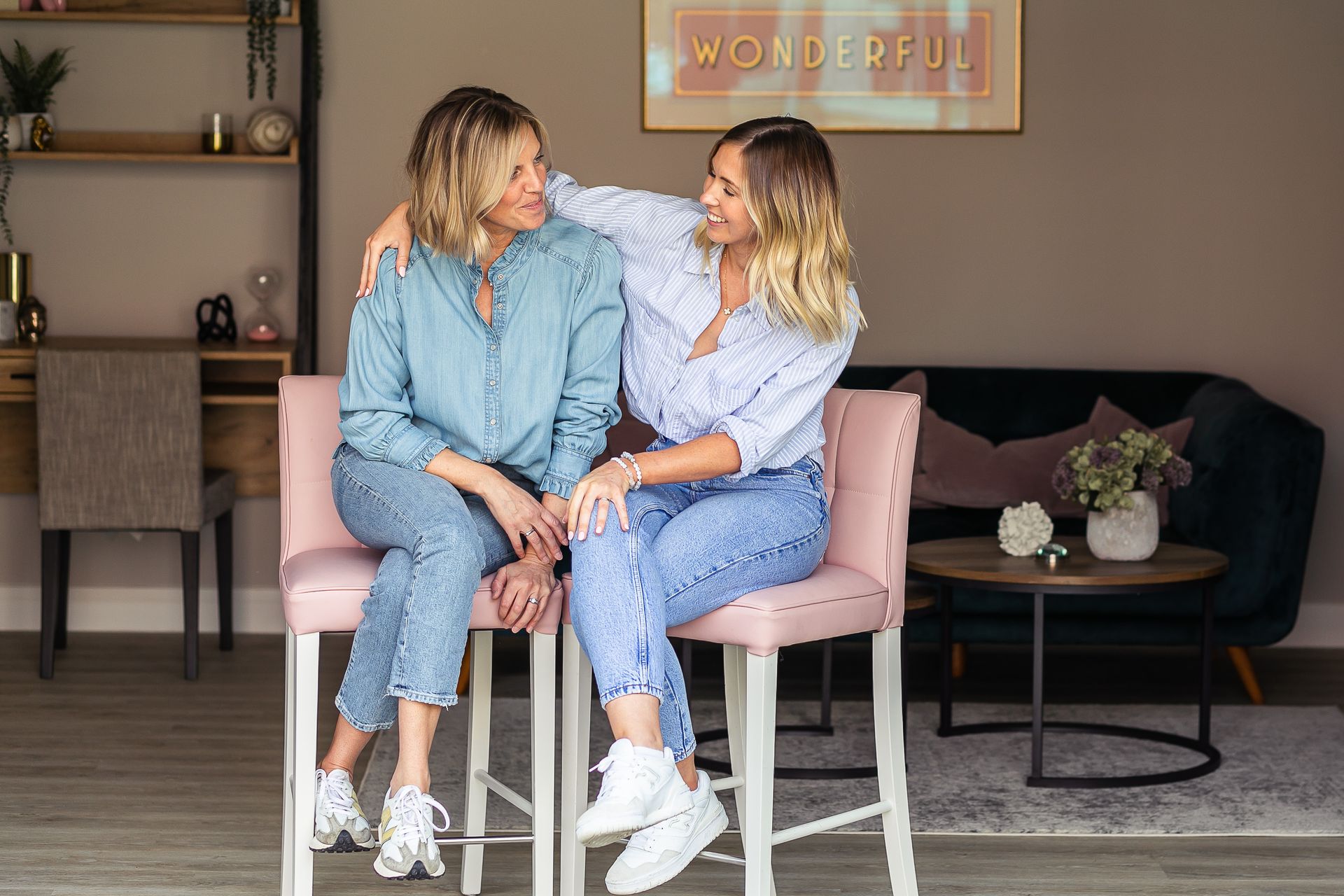 Two people smiling, sitting on pink stools, arm around each other, indoors, modern decor,