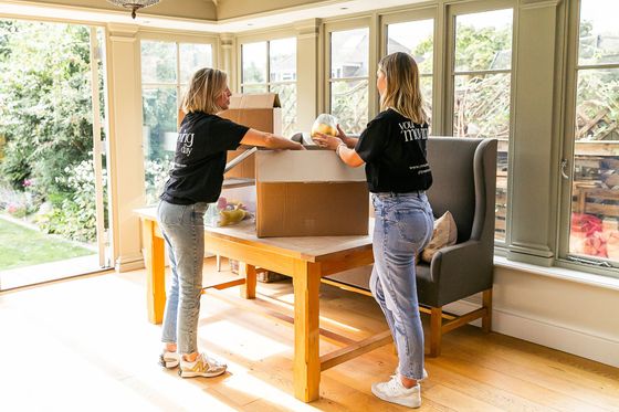 Two people packing items into a cardboard box on a table in a sunroom.