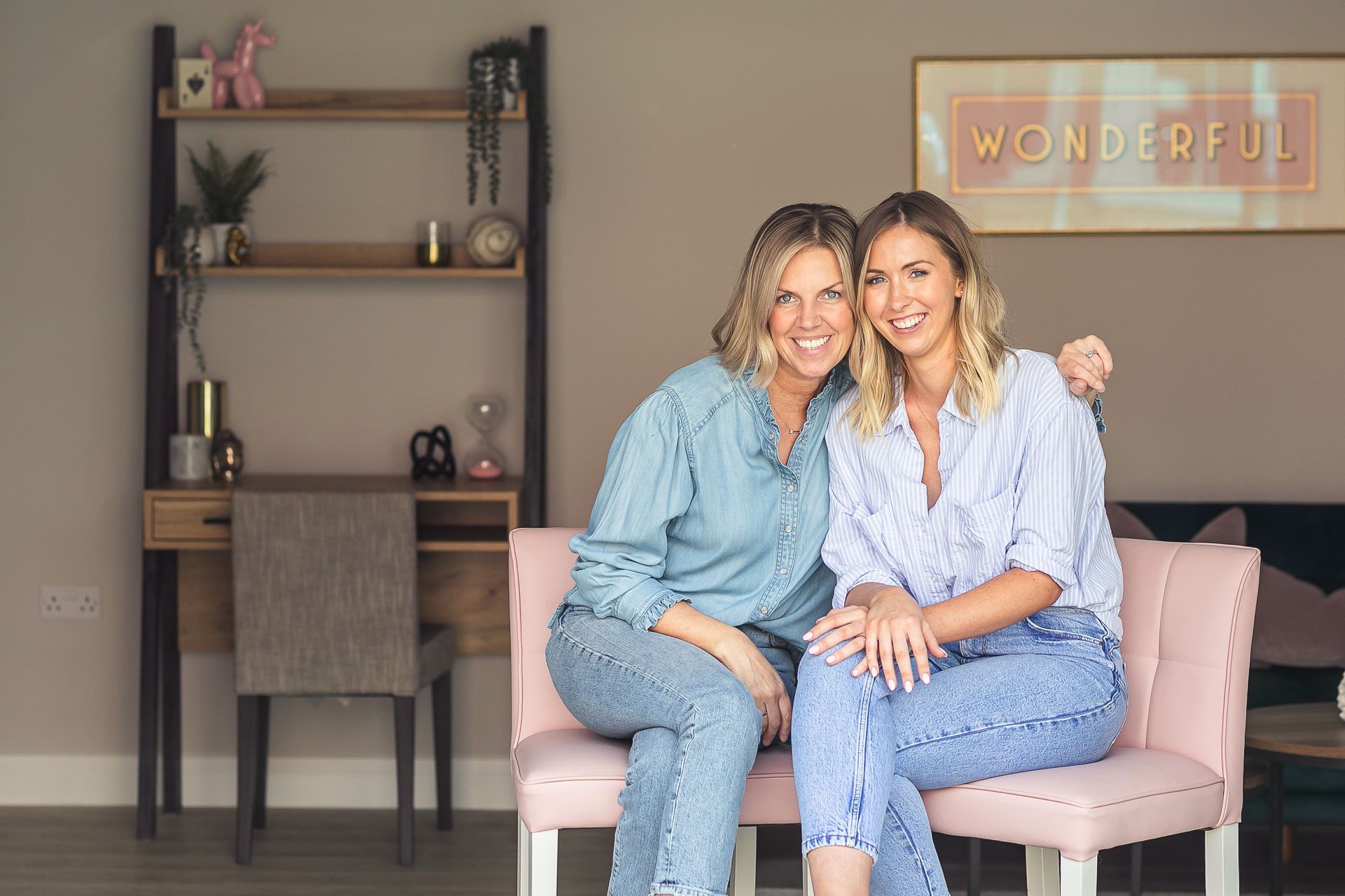 Two women smiling, sitting on a pink chair. Light blue shirts, jeans. Neutral-toned room with desk, shelf, and