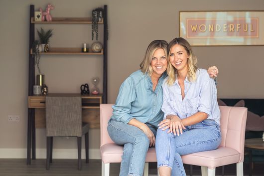 Two smiling women sit on a pink bench, arm around each other, near a desk and decor in a room.
