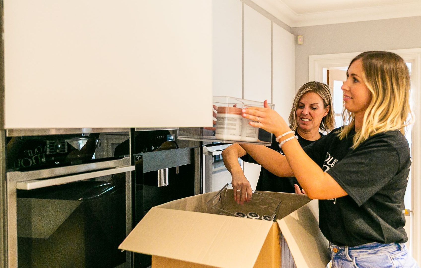 Two women unpacking a box in a kitchen, one holding containers, the other looking on.