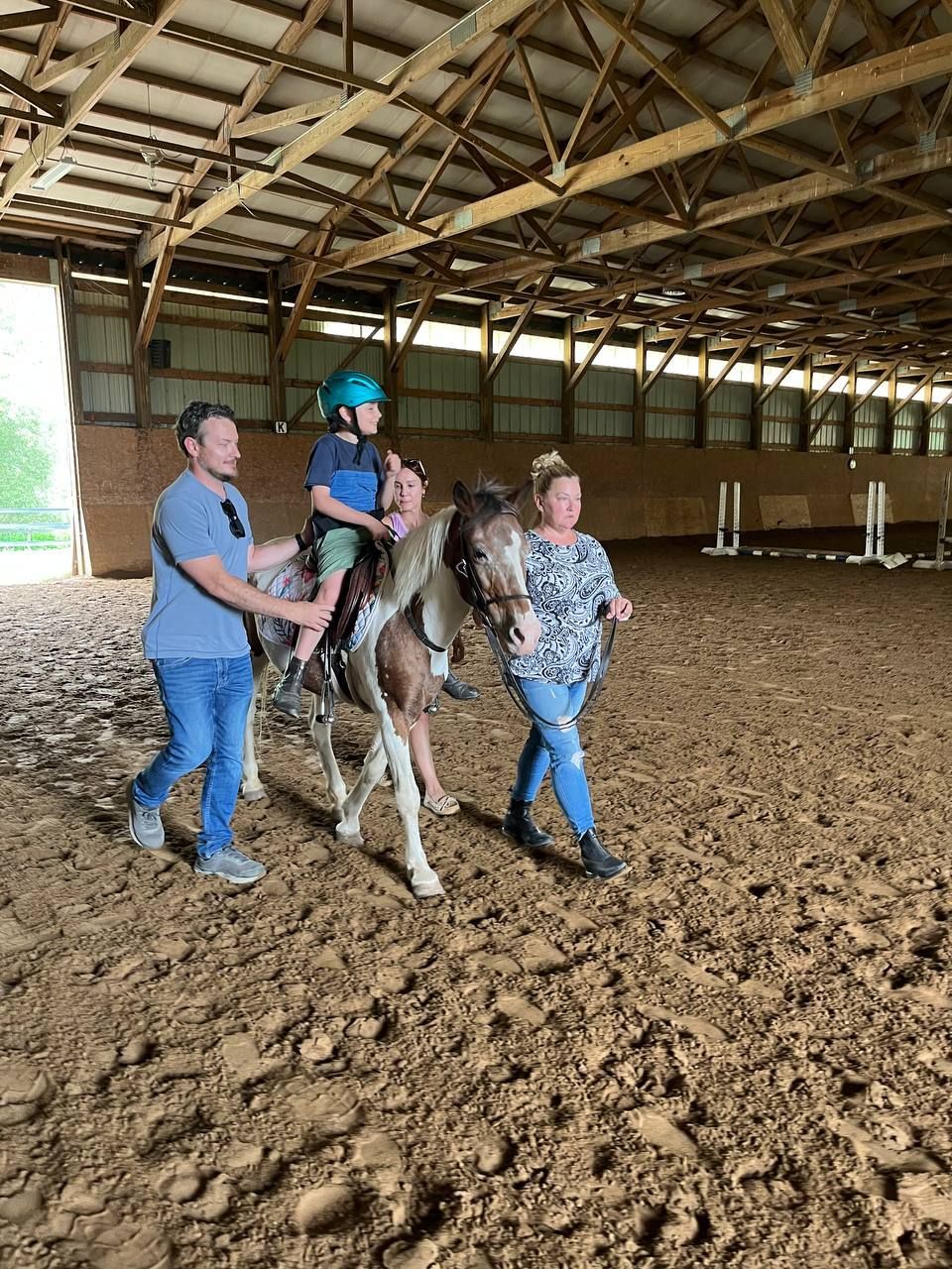 Two adults lead a child on horseback in an indoor riding arena with a dirt floor during an adaptive riding therapy session.