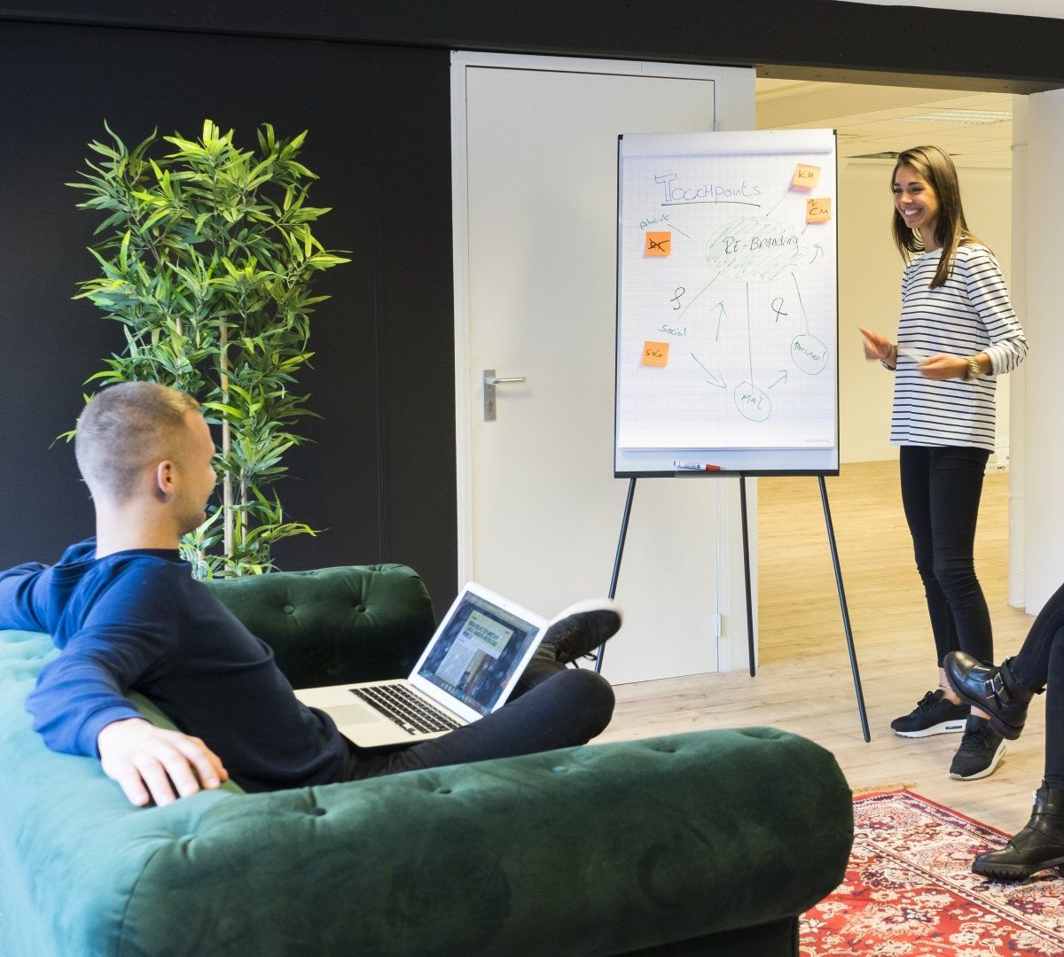 A man is sitting on a couch with a laptop and a woman is standing in front of a whiteboard.