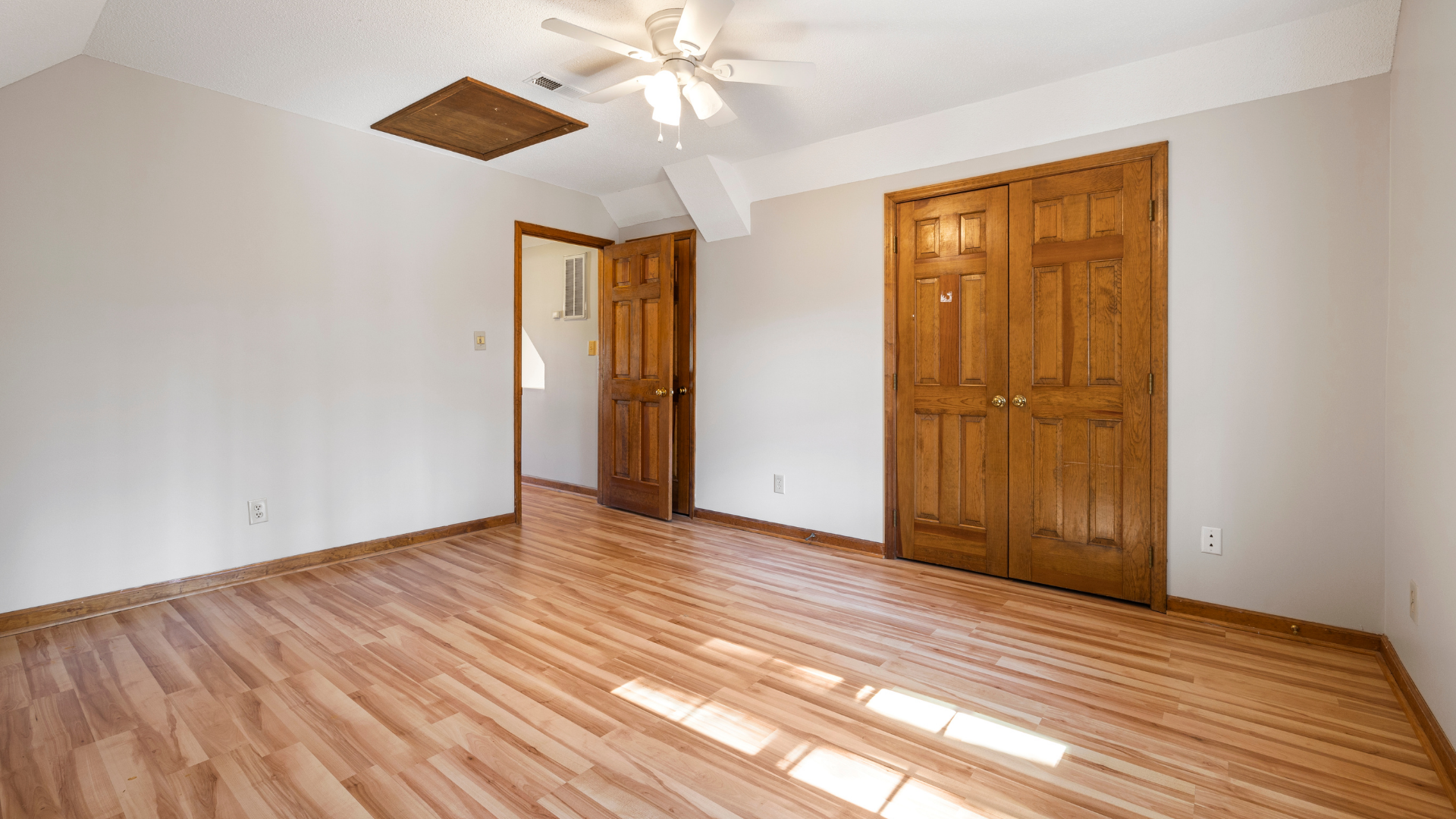 Empty bedroom with hardwood floors, white walls, ceiling fan, and wooden closet doors in bright daylight