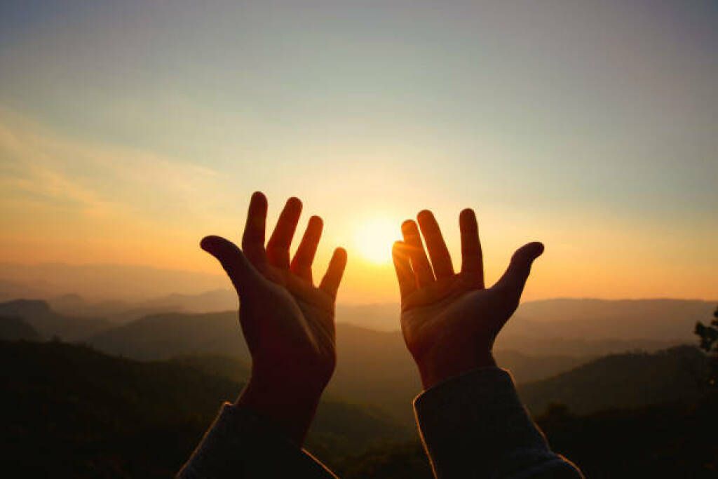 Hands raised toward a setting sun over distant mountains