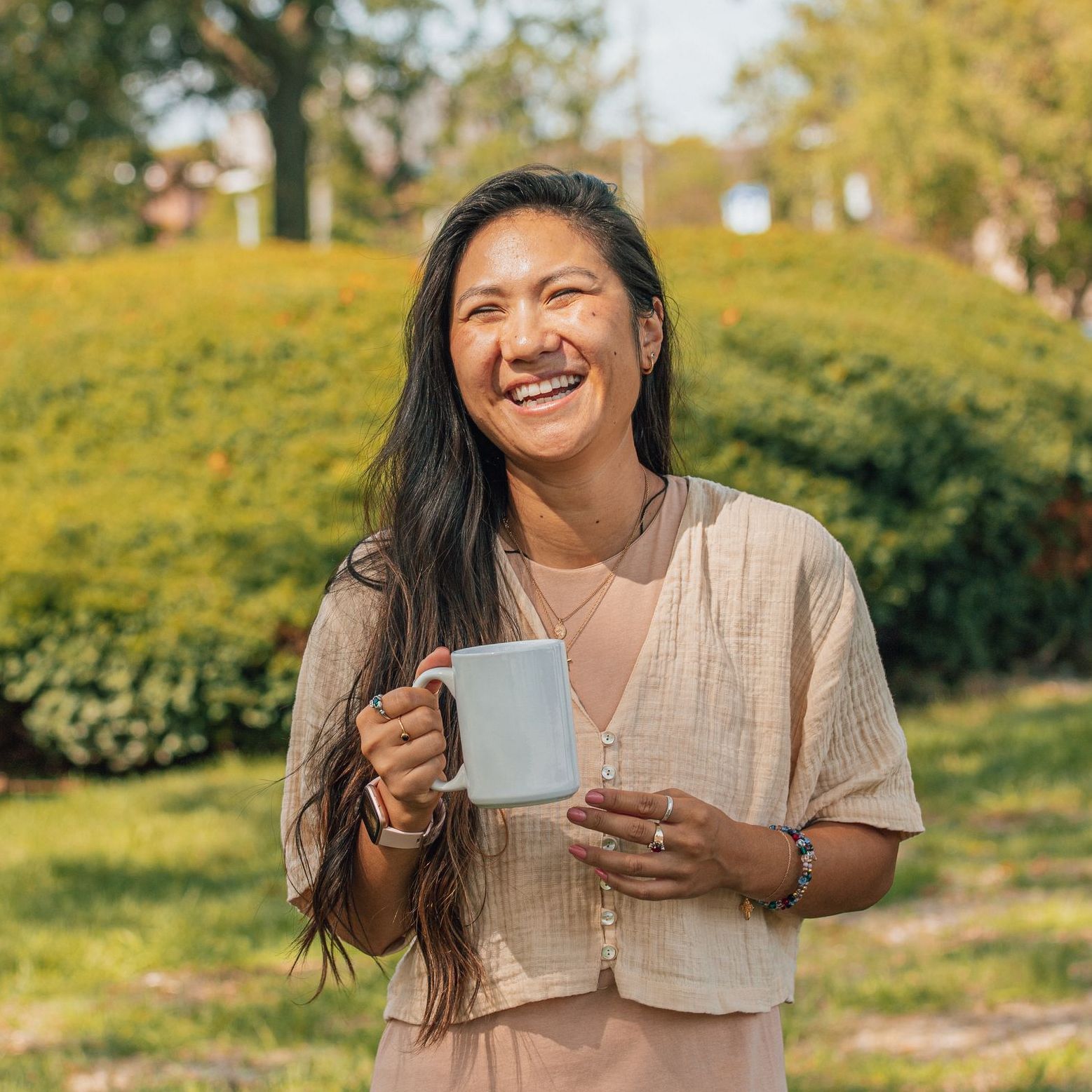 Smiling person holding a mug outdoors in a sunny garden with green bushes