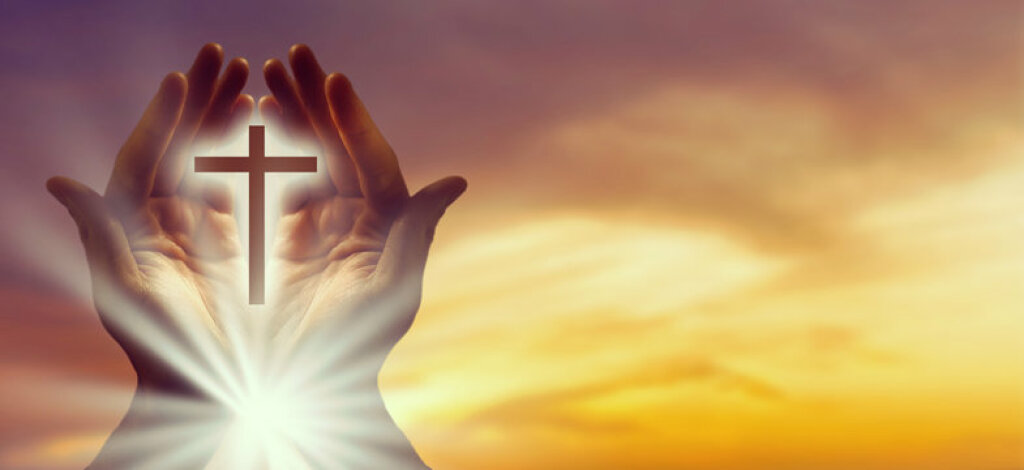 Hands holding a glowing cross against a warm sunset sky